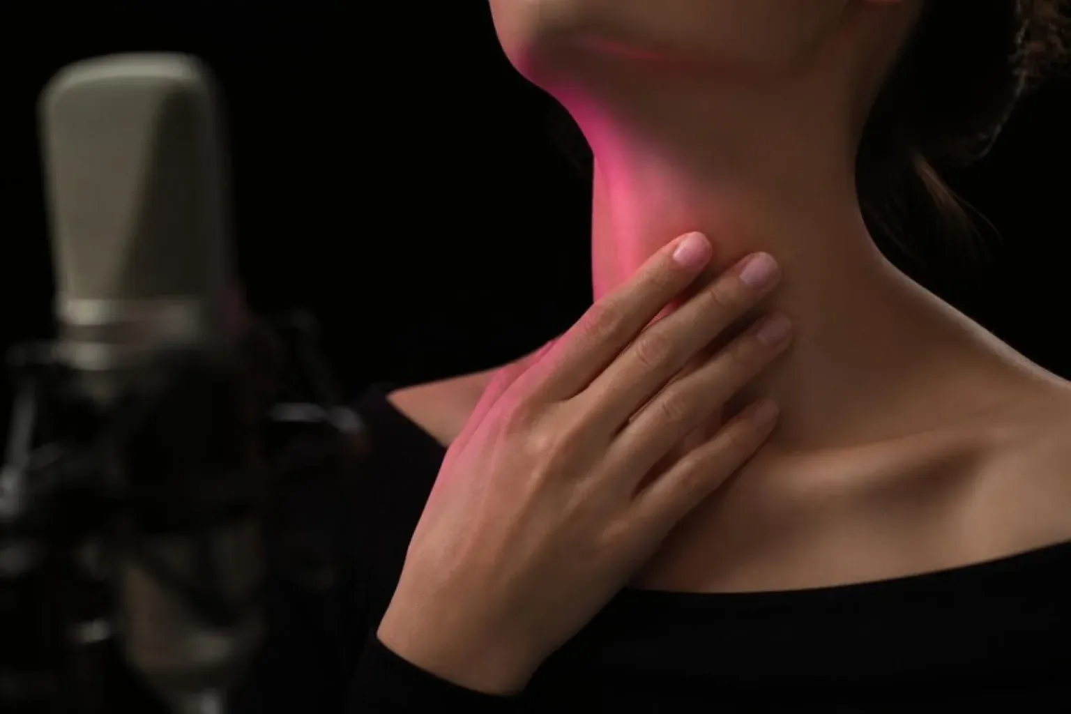 A hand resting on a throat lit by a pink glow, with a blurred studio microphone against a black background