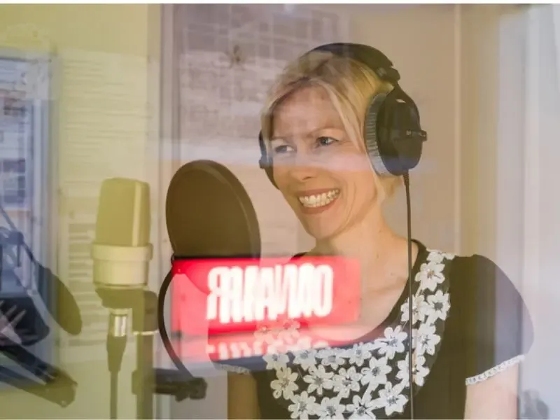 Katie Flamman, comédienne voix off anglaise, souriante au micro dans son studio, casque sur les oreilles, enseigne On Air allumée