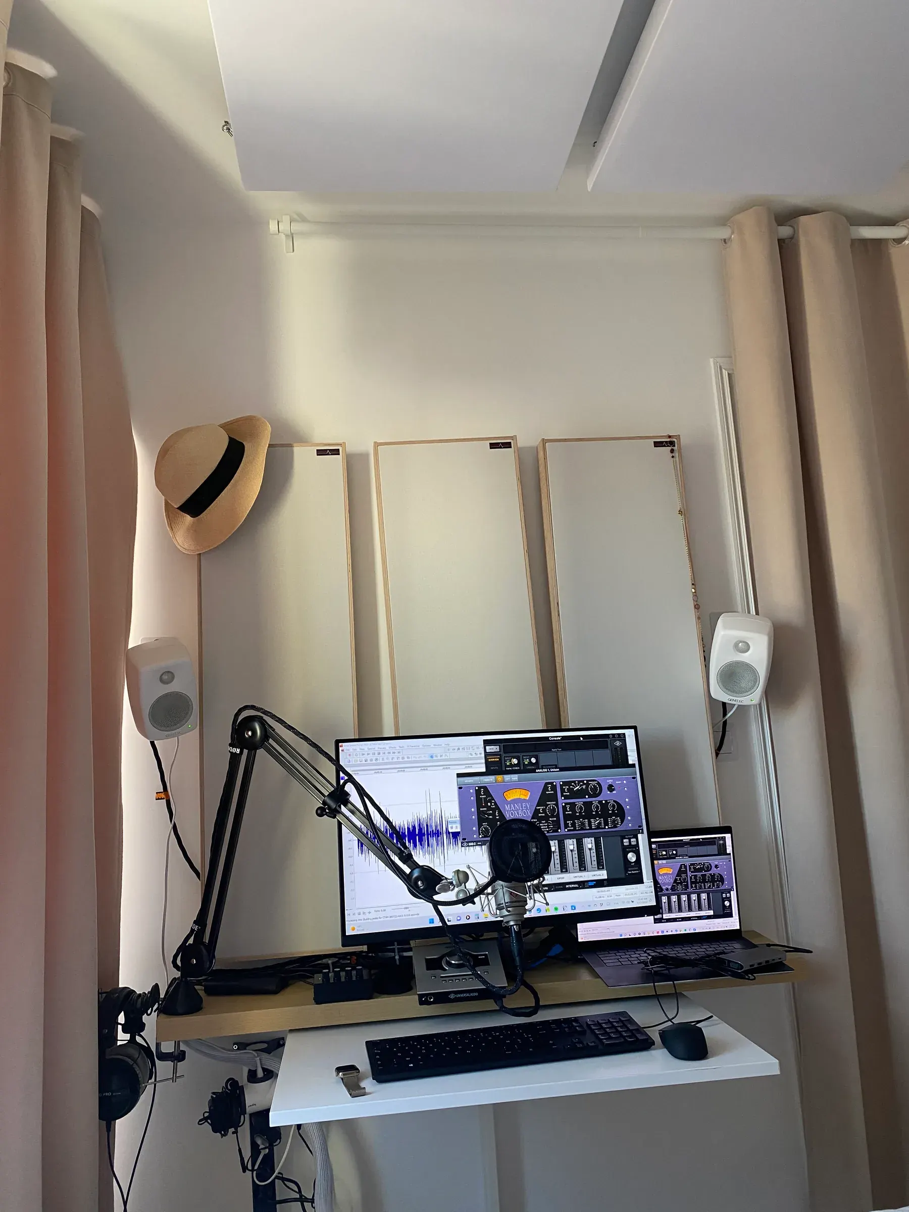 Studio Bis, with pale curtains, acoustic panels on the walls and ceiling, a hanging straw hat, two small white monitors and a microphone on a boom arm in front of a screen and laptop
