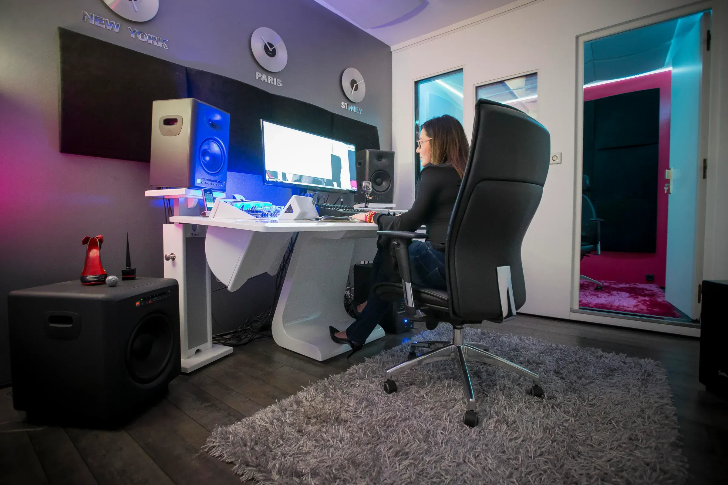 Main studio control room, with a black chair in the foreground, a subwoofer on the left, Estelle Hubert in profile at her desk and the open booth on the right