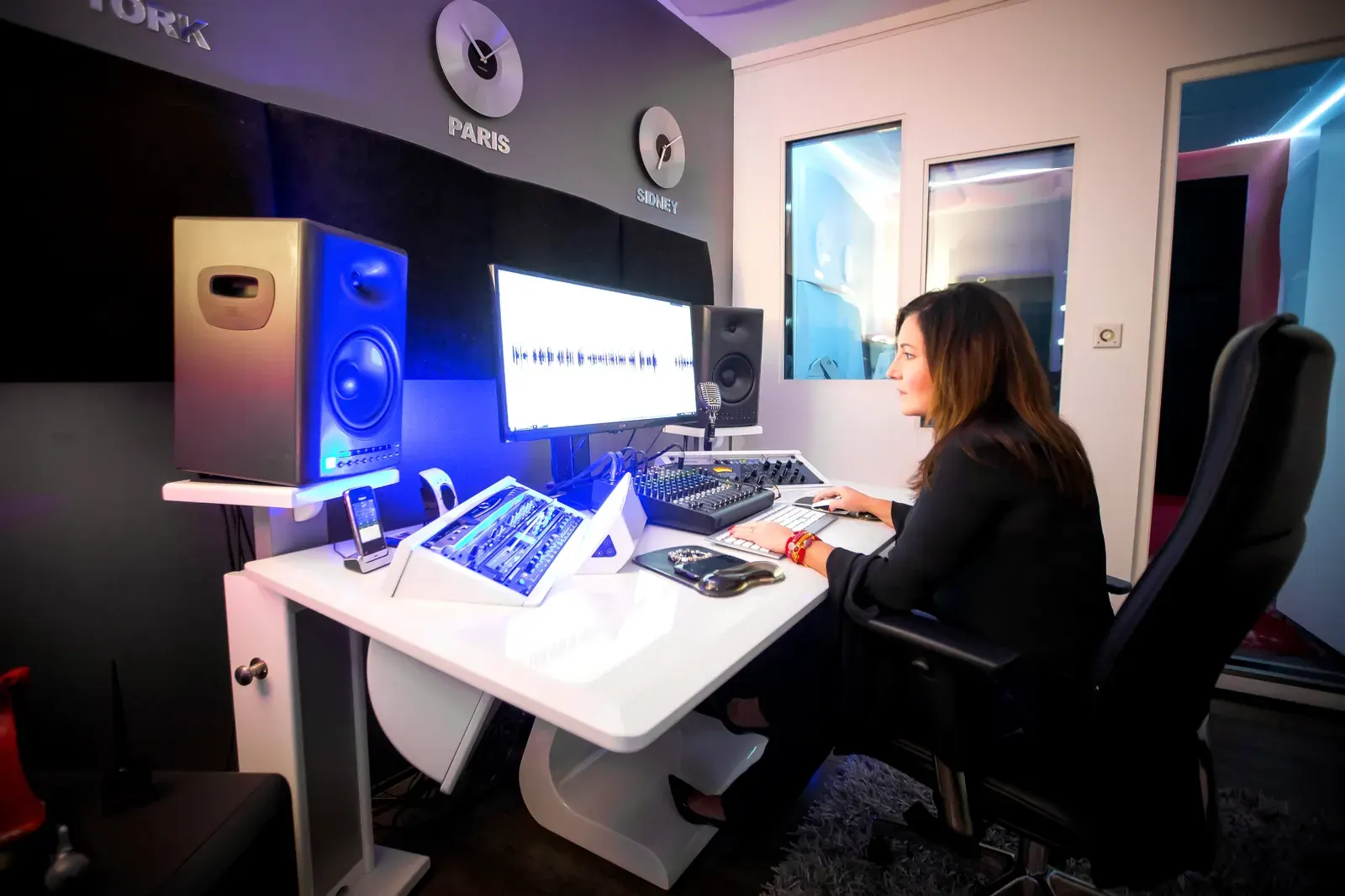 Estelle Hubert, comédienne voix off, dans la control room de son home studio devant ses écrans et ses enceintes de monitoring, la cabine visible derrière la vitre