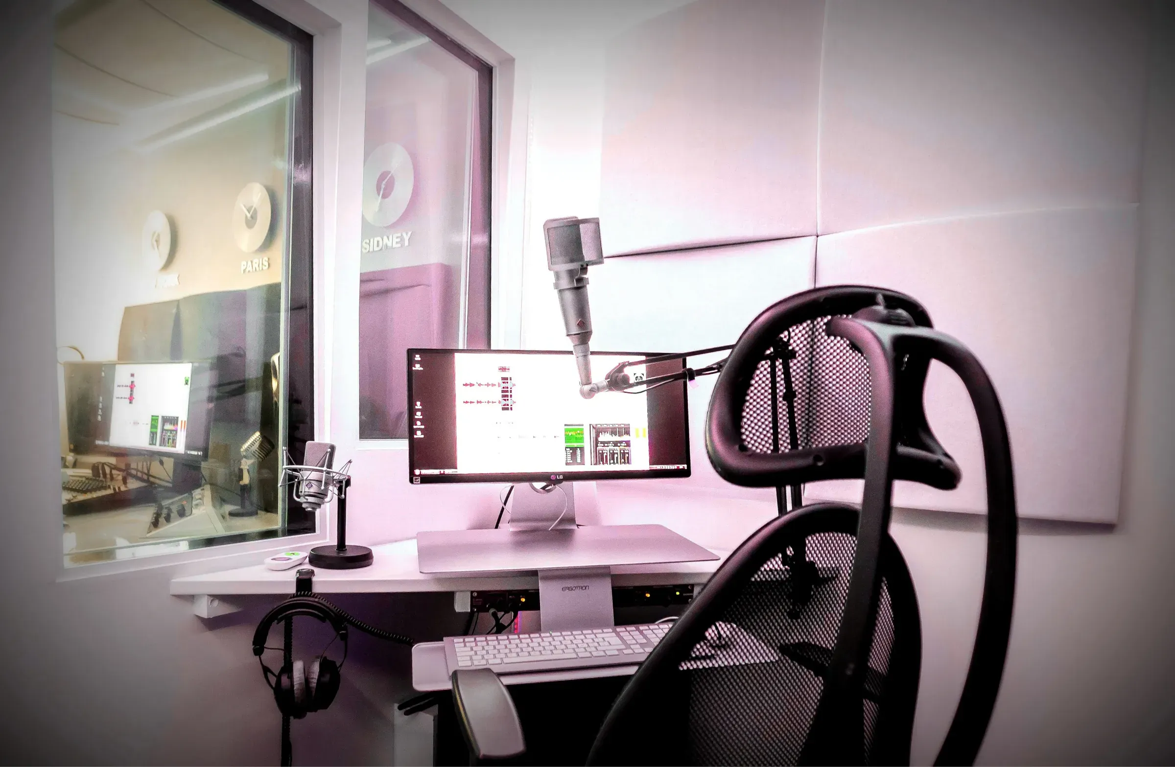 Main studio booth in pink lighting, with a black mesh chair, central screen, white keyboard, headphones and microphone. The control room window is on the left.